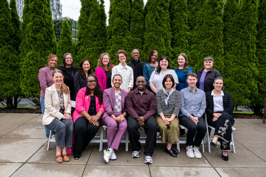GEO staff pose for a group photo outdoors during the 2026 National Conference in Boston, standing and seated in front of tall evergreen trees.