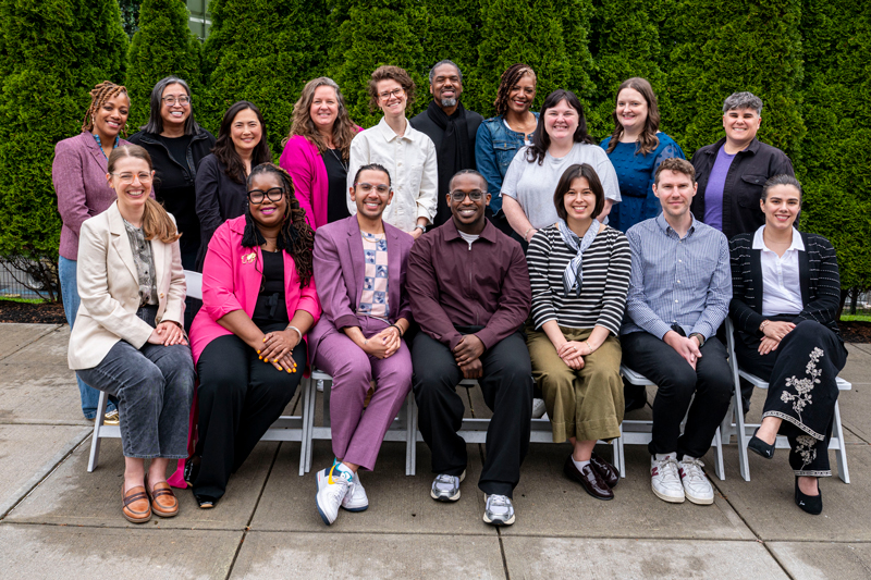 GEO staff pose for a group photo outdoors during the 2026 National Conference in Boston, standing and seated in front of tall evergreen trees.